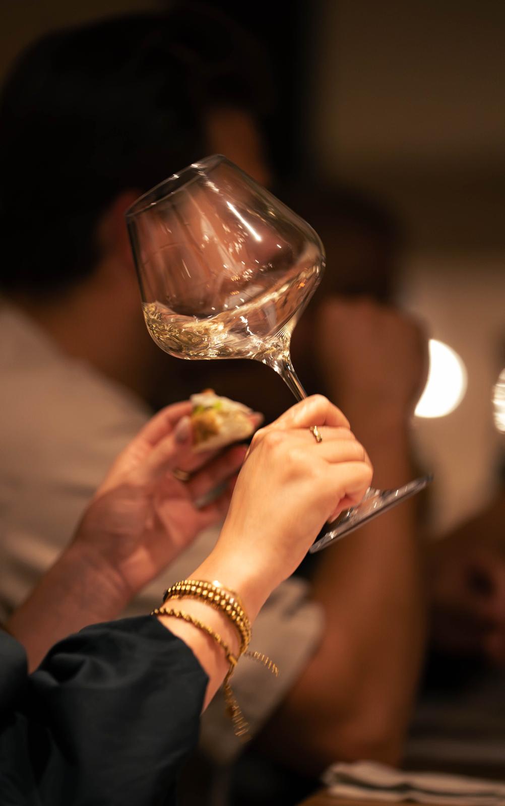Een hand heft een glas witte wijn, met een hapje in de andere hand