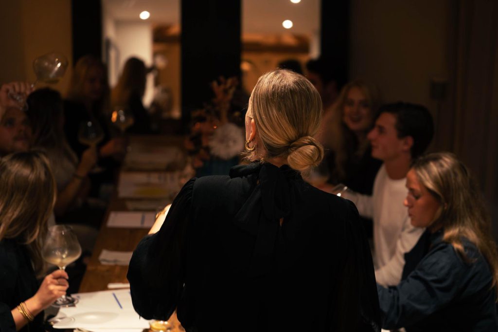 Gasten aan een lange tafel met wijnglazen, de gastvrouw vertelt
